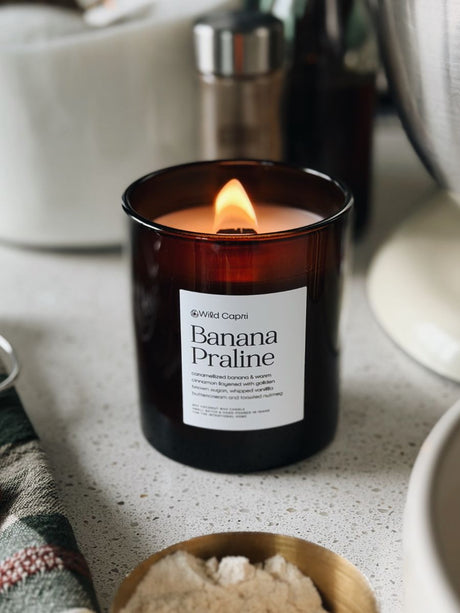 A lit Wild Capri Banana Praline scented candle in an amber glass jar, set on a speckled countertop with kitchen items blurred in the background.