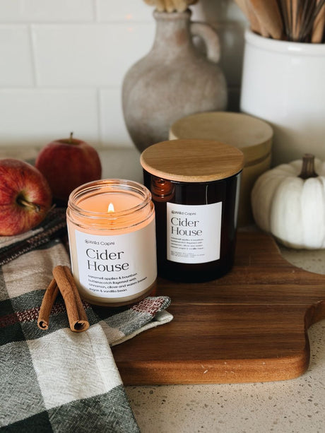 Two Cider House candles from the brand Wild Capri are styled on a wooden cutting board with fall-themed props including apples, cinnamon sticks, a plaid napkin, and a small white pumpkin.