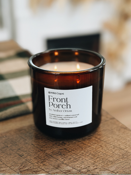 A lit amber glass jar candle labeled 'Front Porch' by Wild Capri, resting on a rustic wooden surface with a softly blurred indoor background.