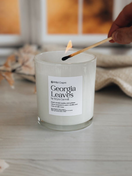 A lit scented candle in a clear glass jar sits on a light wood surface, with a hand holding a match above the flame. The label reads Wild Capri and Georgia Leaves by Eryn Carroll.