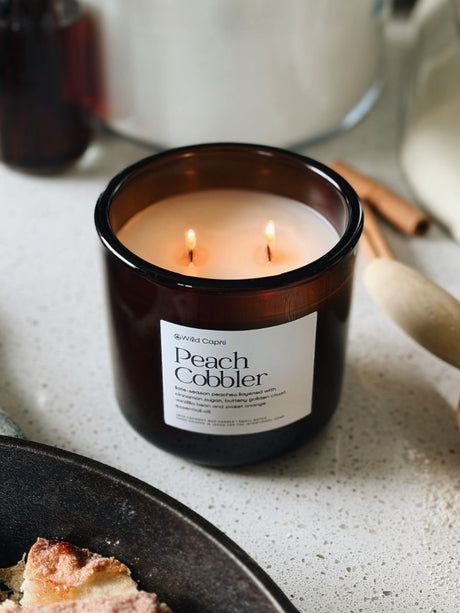 A lit amber glass candle jar labeled 'Peach Cobbler' by Wild Capri, set on a light speckled countertop with warm kitchen props in the background.